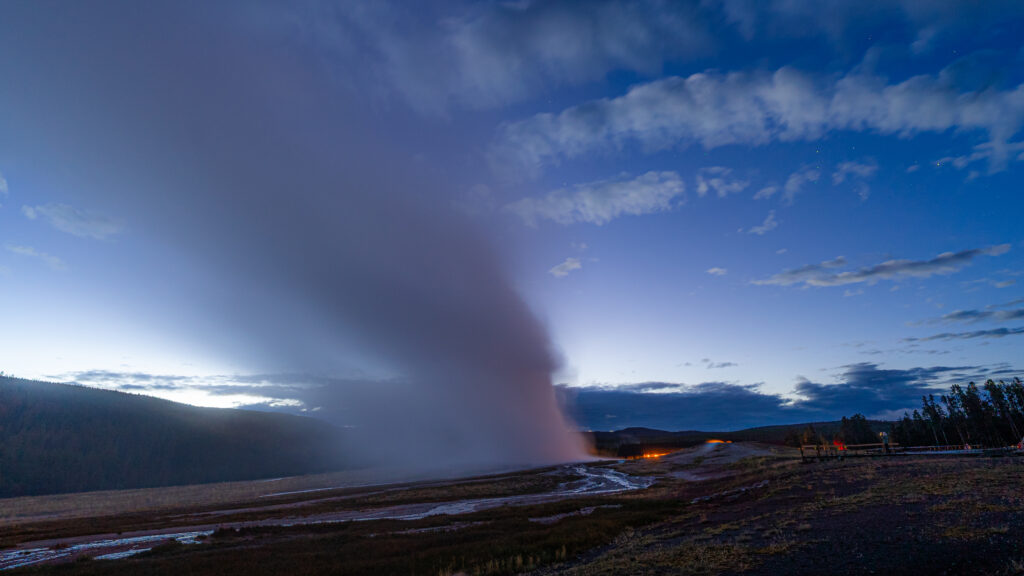 Old Faithful erupting at dawn, steam column bending across the sky, last light on the horizon