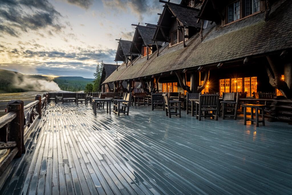 Old Faithful Inn porch at dawn, wet boards reflecting the light, steam rising in the background